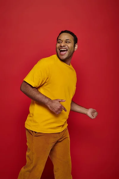 Excité homme indien en vêtements décontractés lumineux souriant et regardant loin sur fond rouge en studio — Photo de stock