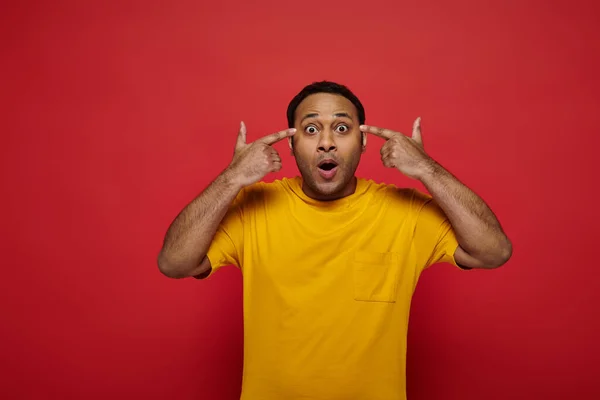 Kopfschüttelnder, schockierter indischer Mann mit Fingern in der Nähe des Kopfes, der auf rotem Hintergrund im Studio in die Kamera blickt — Stockfoto