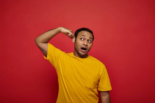 Curious indian man in bright casual clothes looking away and touching ear on red background — Stock Photo