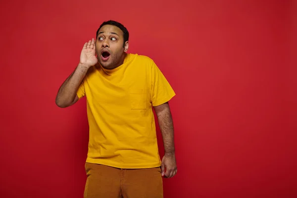 Curious indian man in bright clothes looking away and listening on red backdrop, hand near ear — Stock Photo