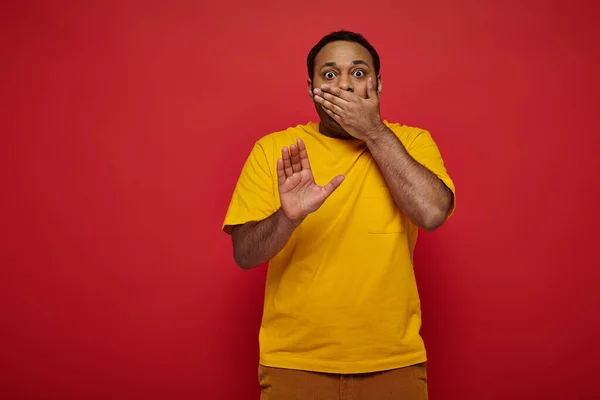 Schockierter indischer Mann in heller Kleidung, der den Mund bedeckt und Stoppschild auf rotem Hintergrund zeigt — Stockfoto