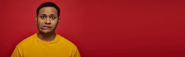 Worried indian man in bright clothes looking at camera and grimacing on red background, banner — Stock Photo