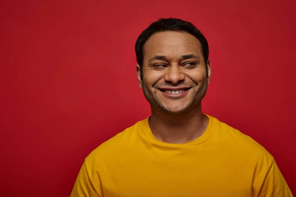 Listige indische Mann in leuchtend gelben Kleidern wegschauen und lächeln auf rotem Hintergrund, Seitenblick — Stockfoto