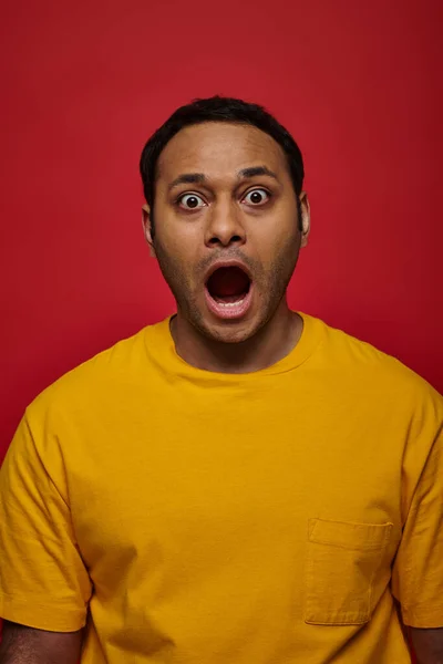 Expresión de choque, hombre indio en ropa amarilla mirando a la cámara con la boca abierta en el telón de fondo rojo - foto de stock