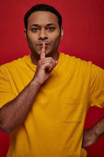Homem indiano em roupas amarelas mostrando sinal de silêncio e olhando para a câmera em pano de fundo vermelho, segredo — Fotografia de Stock