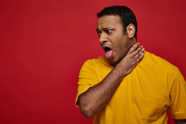 Индийский мужчина в желтой одежде торчащий язык, задыхаясь на красном фоне, затрудненное дыхание — стоковое фото