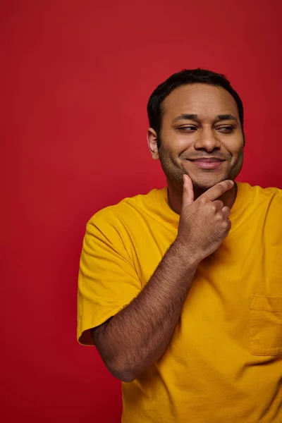 Homem indiano satisfeito em t-shirt amarela tocando queixo e olhando para longe no fundo vermelho, pensativo — Fotografia de Stock