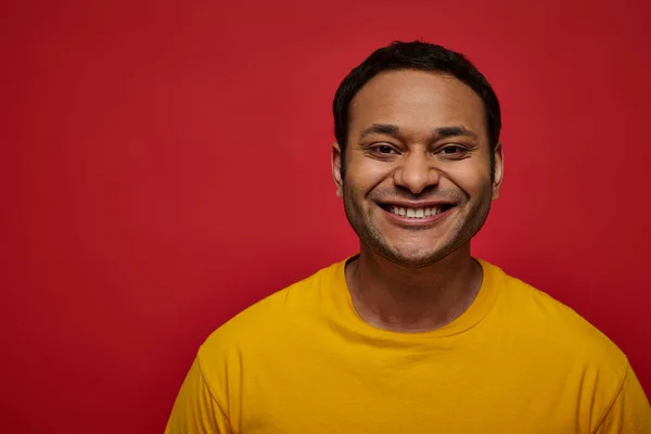 Emoción positiva, hombre indio feliz en camiseta amarilla sonriendo o sonriendo sobre fondo rojo en el estudio - foto de stock