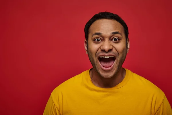 Emoção positiva, homem indiano animado em camiseta amarela com boca aberta no fundo vermelho — Fotografia de Stock