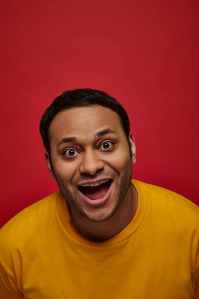 Emoción positiva, hombre indio excitado en camiseta amarilla con la boca abierta sobre fondo rojo, vertical - foto de stock
