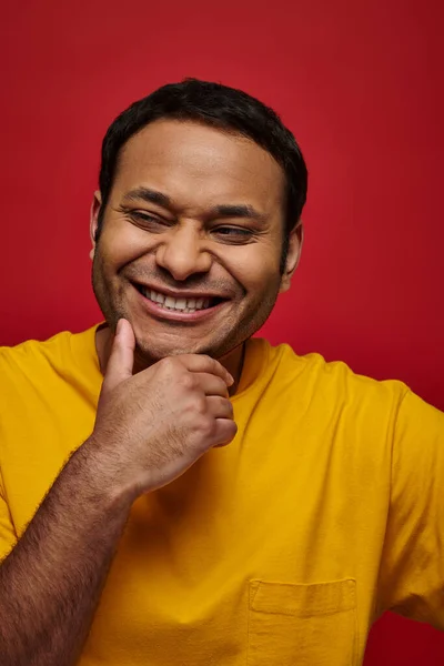 Positive Emotionen, fröhlicher indischer Mann in gelbem T-Shirt lächelnd und berührendes Kinn auf rotem Hintergrund — Stockfoto