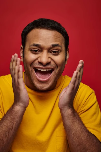 Expresión de la cara, hombre indio excitado en camiseta amarilla gestos sobre fondo rojo, boca abierta - foto de stock