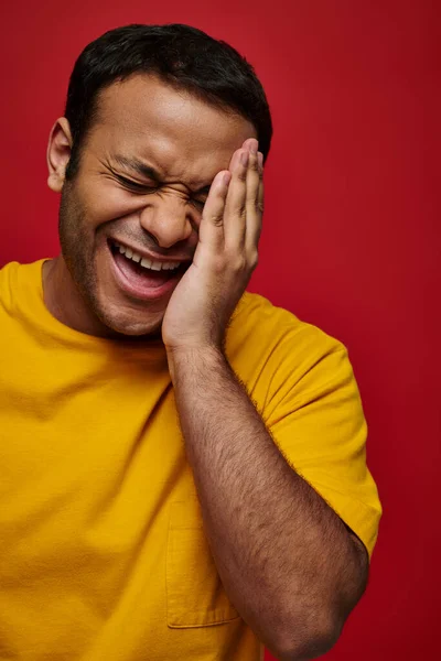 Expression du visage, homme indien embarrassé en t-shirt jaune riant et touchant visage sur fond rouge — Photo de stock
