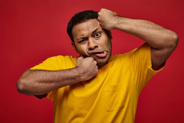 Expressão facial, homem indiano confuso em camiseta amarela perfurando-se no rosto em pano de fundo vermelho — Fotografia de Stock
