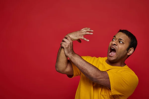 Expresión de la cara, hombre indio asustado en camiseta amarilla perdiendo el control de su mano sobre el fondo rojo - foto de stock