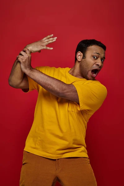 Вираз обличчя, шокований індійський чоловік у жовтій футболці втрачає контроль над рукою на червоному тлі — стокове фото