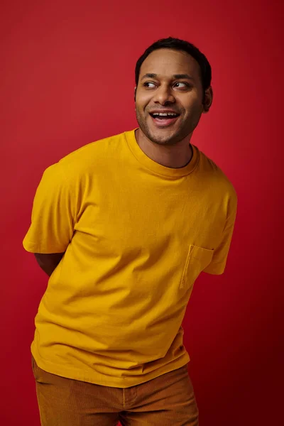 Allegro uomo indiano in t-shirt gialla distogliendo lo sguardo e sorridendo su sfondo rosso, espressione del viso — Foto stock