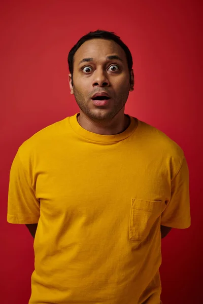 Homme indien étonné en t-shirt jaune regardant la caméra sur fond rouge, expression du visage — Photo de stock