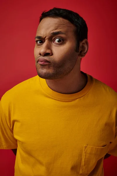 Dudoso hombre indio en camiseta amarilla mirando a la cámara sobre fondo rojo, expresión de la cara - foto de stock
