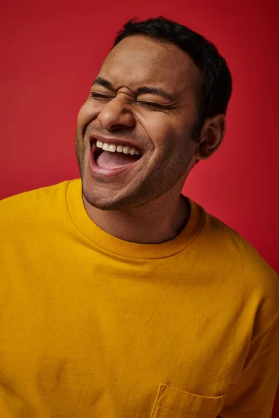 Espressione del viso, uomo indiano eccitato in t-shirt gialla ridere su sfondo rosso, bocca aperta — Foto stock