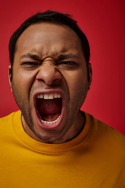 Expresión de la cara, hombre indio emocional en camiseta amarilla gritando sobre fondo rojo, boca abierta - foto de stock