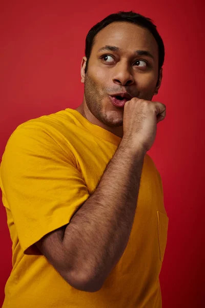 Expression du visage, homme indien en t-shirt jaune montrant la réaction tout en regardant loin sur fond rouge — Photo de stock