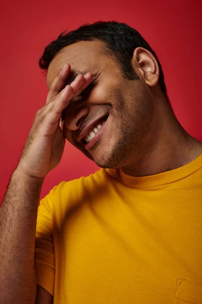 Tímido hombre indio en camiseta amarilla sonriendo y cubriendo los ojos con la mano sobre fondo rojo en el estudio - foto de stock