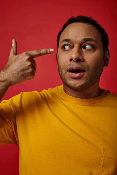 Schockierter indischer Mann in gelbem T-Shirt zeigt mit erhobenem Zeigefinger auf rotem Hintergrund im Studio — Stockfoto