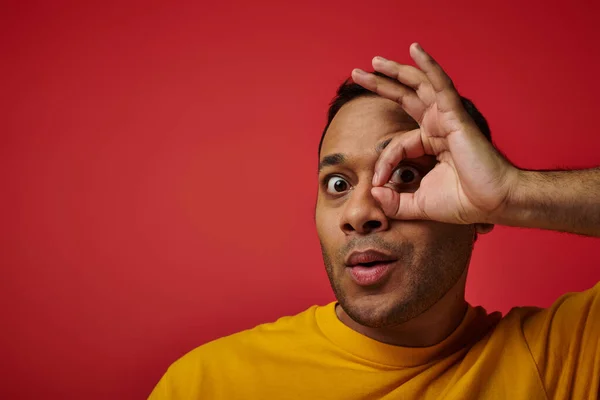 Hombre indio sorprendido en camiseta amarilla que muestra signo de bien, mano cerca de fondo de la cara en el estudio - foto de stock