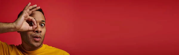 Homme indien étonné en t-shirt jaune montrant signe correct, main près du visage en arrière-plan en studio, bannière — Photo de stock