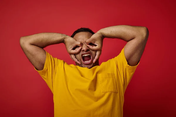 Uomo indiano emotivo in t-shirt gialla urlando e gesticolando su sfondo rosso, viso espressivo — Foto stock