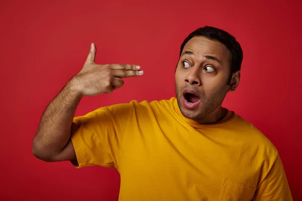 Homem indiano chocado em t-shirt amarela gesticulando em fundo vermelho, apontando com os dedos perto da cabeça — Fotografia de Stock