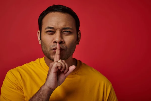 Indian man in yellow t-shirt showing shh gesture on red backdrop, secret, finger near lips — Stock Photo