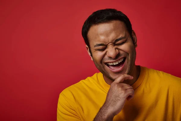 Joyous indian man in yellow t-shirt smiling with closed eyes on red backdrop in studio, portrait — Stock Photo