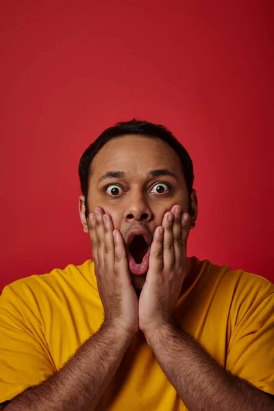 Homme indien étonné en t-shirt jaune regardant la caméra avec les yeux grands ouverts sur fond rouge — Photo de stock