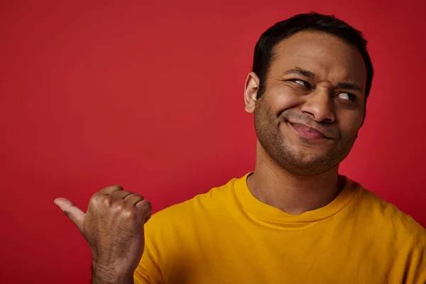 Hombre indio escéptico en camiseta amarilla apuntando con el pulgar y mirando hacia otro lado en el fondo rojo - foto de stock