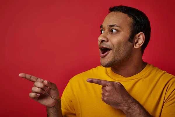 Homme indien étonné en t-shirt jaune pointant avec les doigts et regardant loin sur fond rouge — Photo de stock