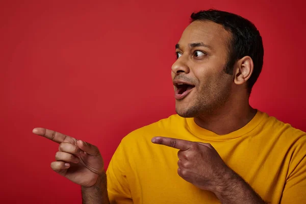 Astonished indian man in yellow t-shirt pointing with fingers and looking away on red backdrop — Stock Photo