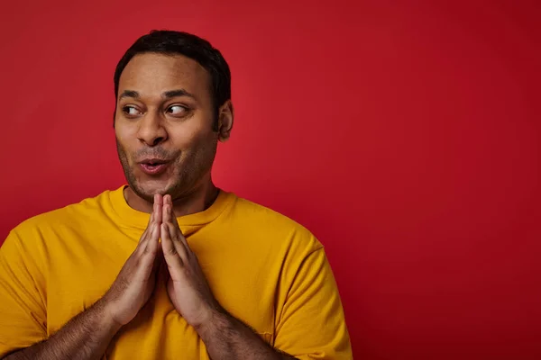 Astuto hombre indio en camiseta amarilla agarrándose de las manos y planeando algo sobre fondo rojo, cara astuta - foto de stock