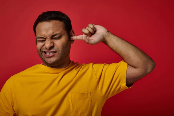 Hombre indio en camiseta amarilla cerrando los ojos y recogiendo su oreja con el dedo en el fondo rojo, mueca - foto de stock