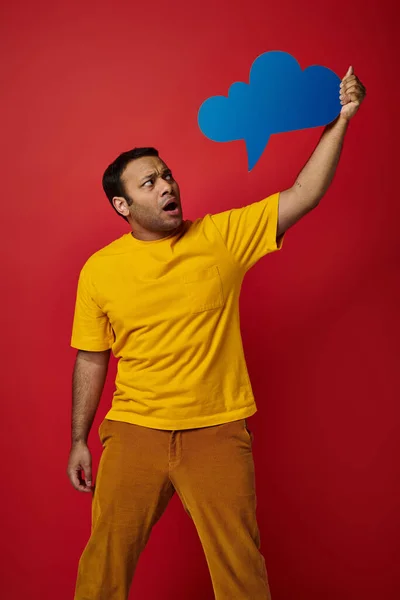 Homme indien choqué en t-shirt jaune tenant bulle de pensée vierge sur fond rouge, visage étonné — Photo de stock