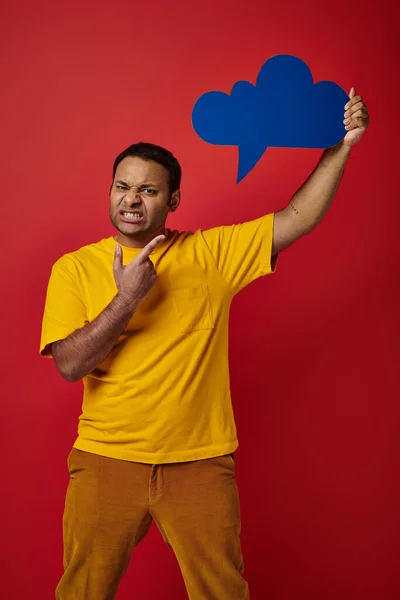 Homme indien mécontent en t-shirt jaune pointant bulle de pensée vierge sur fond rouge, grimace visage — Photo de stock