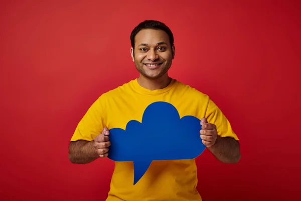 Cheerful indian man in yellow t-shirt holding blue blank thought bubble on red backdrop, happy face — Stock Photo