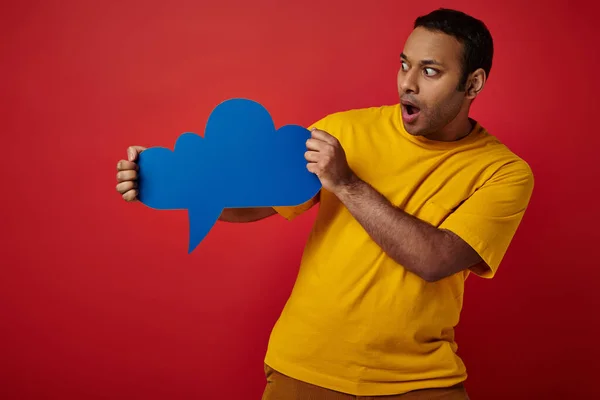 Homem indiano espantado em t-shirt amarela olhando em branco pensamento bolha no fundo vermelho, rosto chocado — Fotografia de Stock
