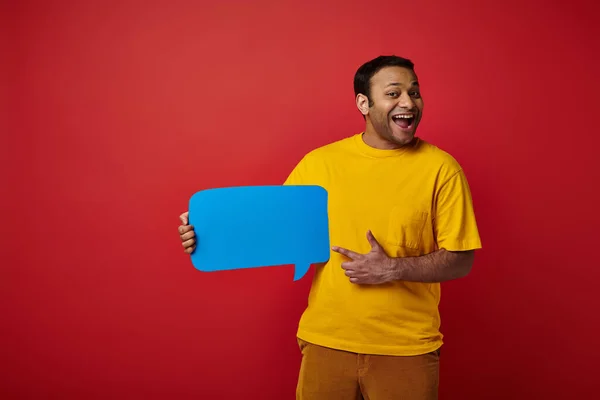 Homem animado em camiseta amarela apontando com o dedo para a bolha de fala em branco no fundo vermelho, feliz — Fotografia de Stock