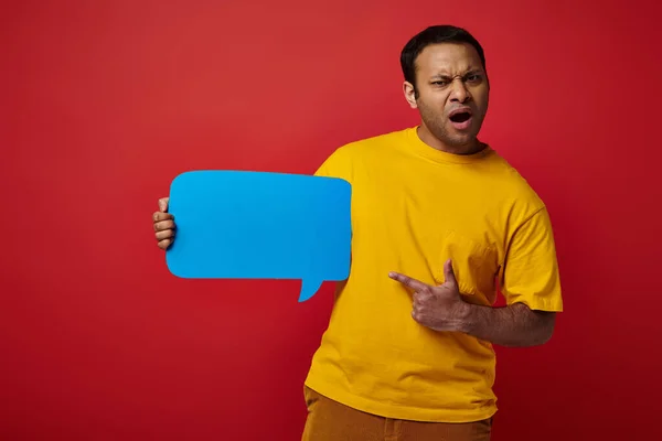 Homem chocado em camiseta amarela apontando para bolha de fala em branco no fundo vermelho, descontente — Fotografia de Stock