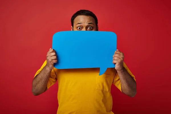 Indian man with eyes wide open hiding behind blank speech bubble on red background, emotional — Stock Photo