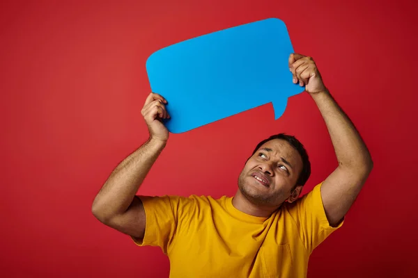 Displeased indian man in yellow t-shirt holding blank speech bubble above head on red background — Stock Photo