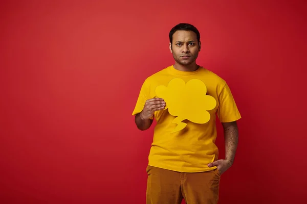 Angespannter indischer Mann hält leere Sprechblase und steht mit der Hand in der Tasche auf rotem Hintergrund — Stockfoto