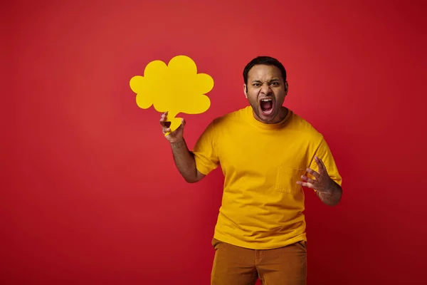 Angry indian man in yellow t-shirt holding blank speech bubble and screaming on red background — Stock Photo